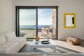 Ein modernes Wohnzimmer mit einem weißen Sofa, einem Couchtisch mit Dekoration, einer großen Schiebeglasdoor, die auf einen Balkon mit Meerblick führt, und einem gelb gerahmten Spiegel an der Wand.