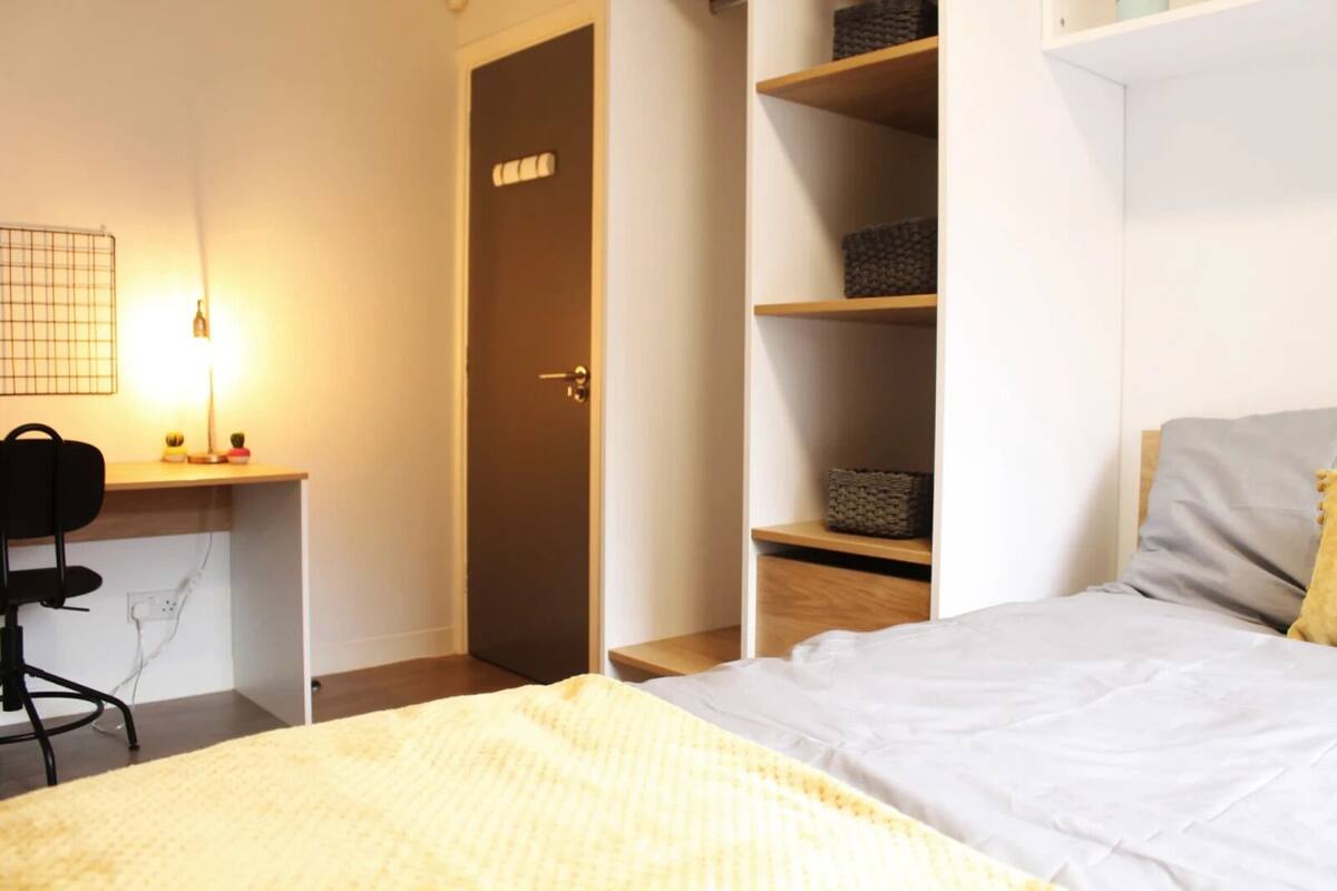 A bedroom with a bed, a desk with a chair, a wardrobe, and shelves with baskets.