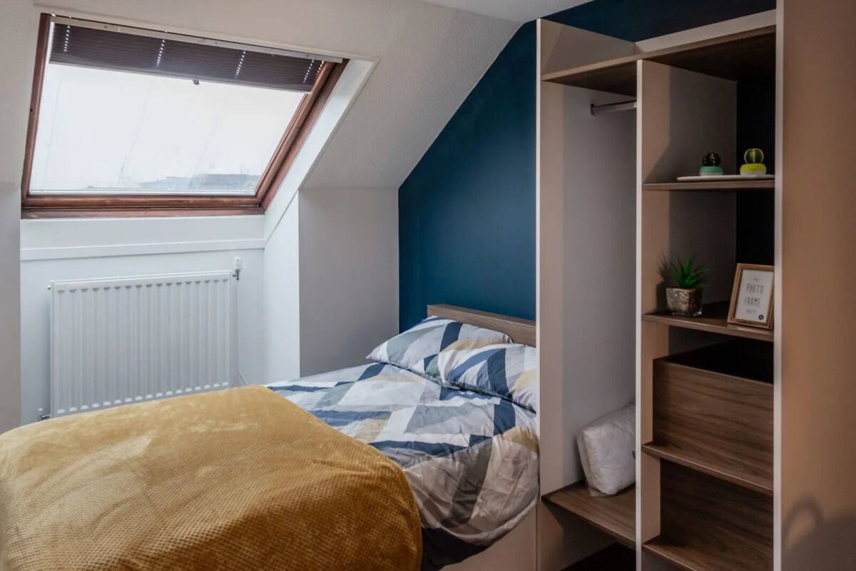 A bedroom with a sloped ceiling, a window with blinds, a radiator, a bed with a patterned bedspread, a wooden bench, and a built-in shelving unit with a plant and a photo frame.