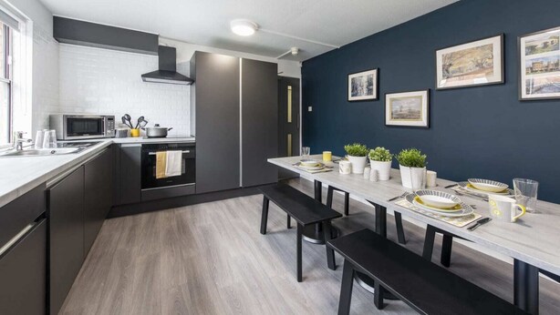 A modern kitchen with a dining area, featuring a large island, dark cabinetry, and a blue accent wall. - For Students Only Regents Court in Leicester (Leicester)