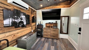Une pièce avec un plafond en bois, un ventilateur de plafond, un téléviseur fixé au mur, une commode en bois, un lit recouvert d’une couverture verte et un climatiseur mural.