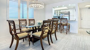 Un comedor con mesa de vidrio, sillas de madera, una araña y una cocina al fondo.