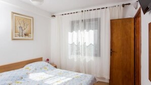 Ein Schlafzimmer mit einem Bett, einer Holztür, einem Fenster mit durchscheinenden Vorhängen und einem gerahmten Bild an der Wand.