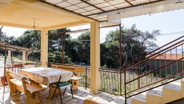 Ein überdachter Essbereich im Freien mit Tisch und Bänken, einer Treppe sowie Blick auf Bäume und ein Gebäude im Hintergrund.