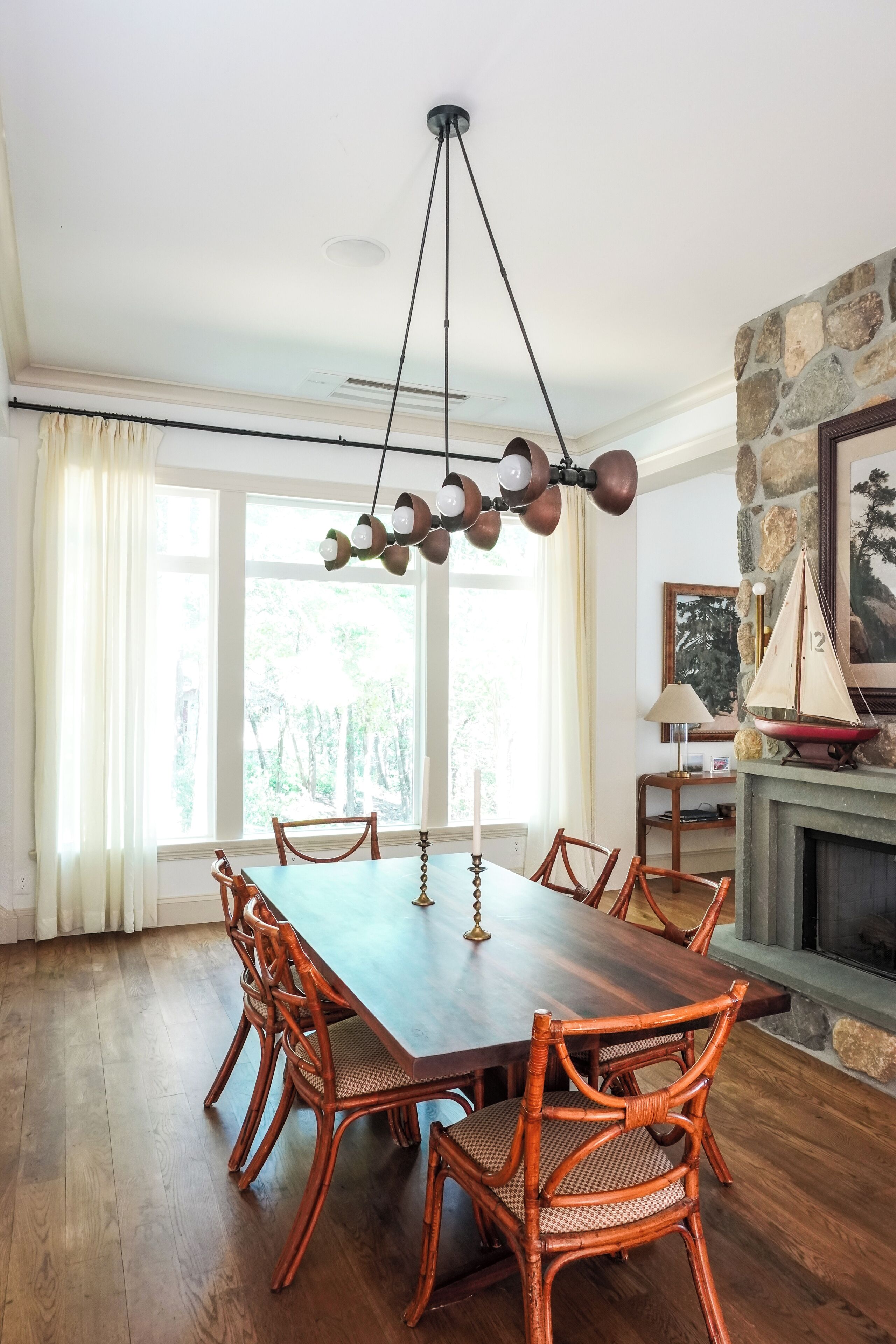 A dining room with a wooden table and wicker chairs, a stone fireplace, and a large window with curtains.