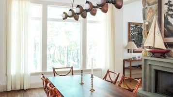 A dining room with a wooden table and wicker chairs, a stone fireplace, and a large window with curtains.