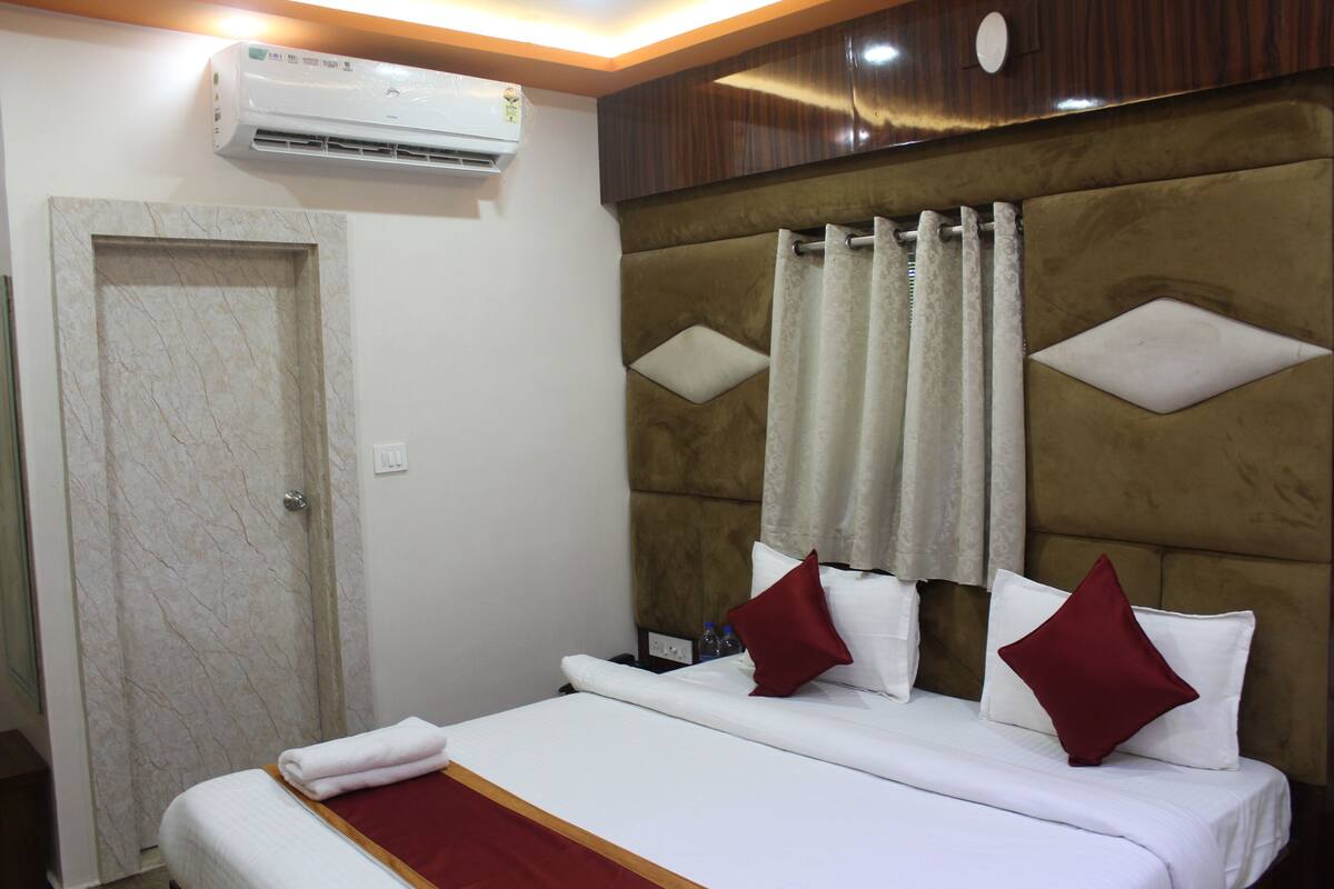 A hotel room with a marble-framed door, a bed with white bedding and red pillows, a headboard with a diamond pattern, and an air conditioning unit above the door.