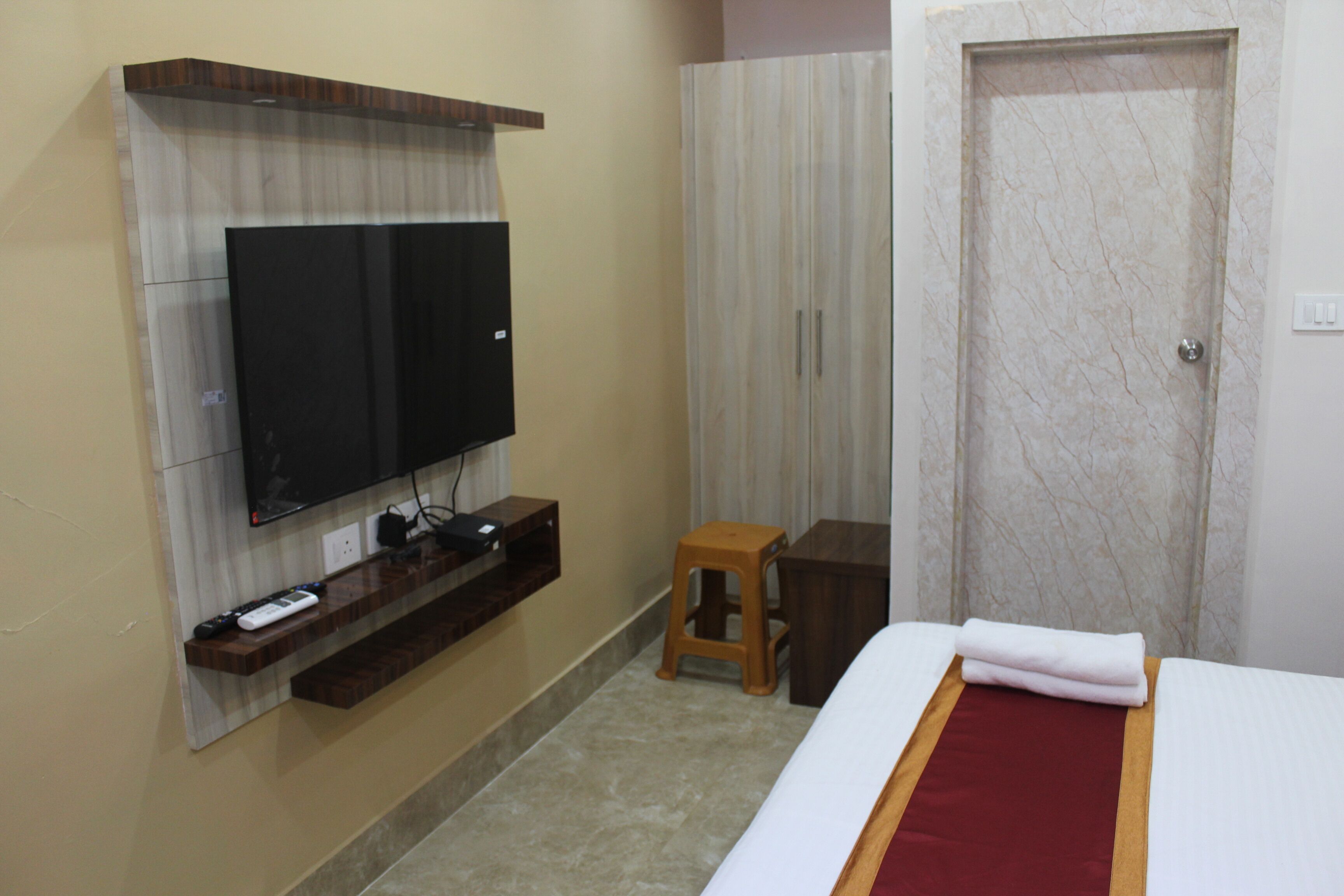 A hotel room with a flat-screen TV mounted on the wall, a wooden nightstand, a small stool, a bed with a red and white bedspread, and a door with a marble-like pattern.