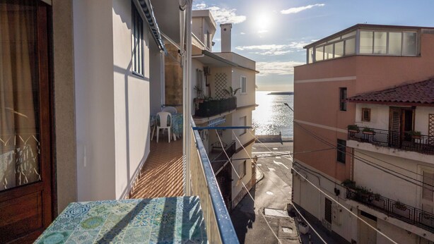A balcony with a view of the sea, a table with a patterned tablecloth, and a chair.