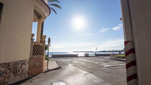 A street view with a clear sky, a body of water in the background, and a building with a stone facade and palm trees.