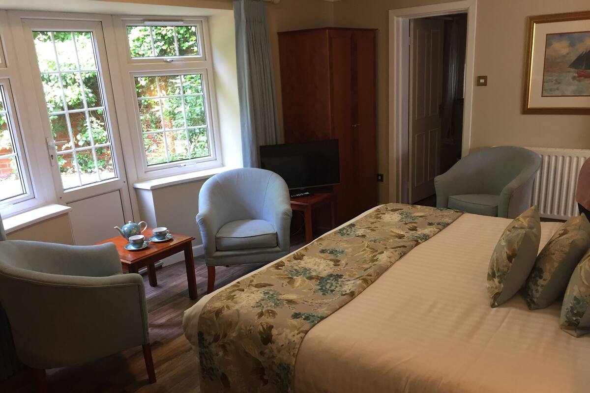 Une chambre d’hôtel avec un lit, deux fauteuils, une télévision et une fenêtre donnant sur un paysage verdoyant.