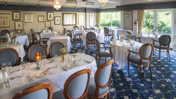 A formal dining room with multiple tables set for service, framed artwork on the walls, and large windows providing natural light.