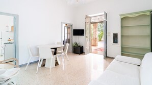 Ein modernes Wohnzimmer mit einem weißen Sofa, einem Esstisch mit Stühlen, einem Fernseher und einer grünen Regaleinheit.