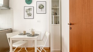 Une petite cuisine avec une table blanche, deux chaises, une horloge et, au mur, des reproductions encadrées de gravures botaniques.