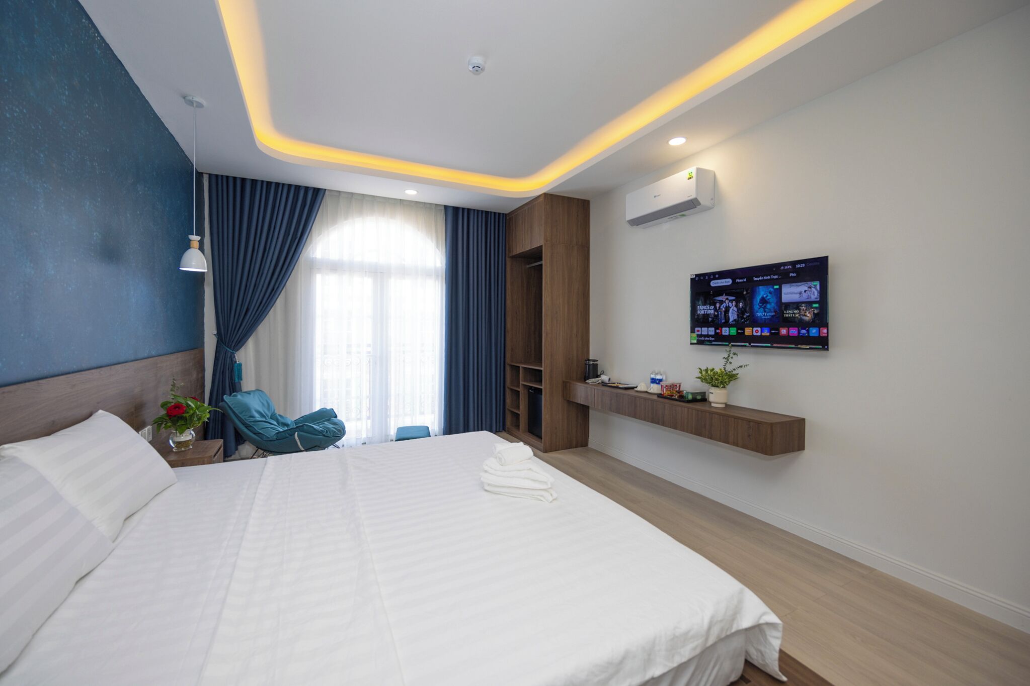 A modern hotel room with a large bed, a TV mounted on the wall, and a wooden shelf with amenities.