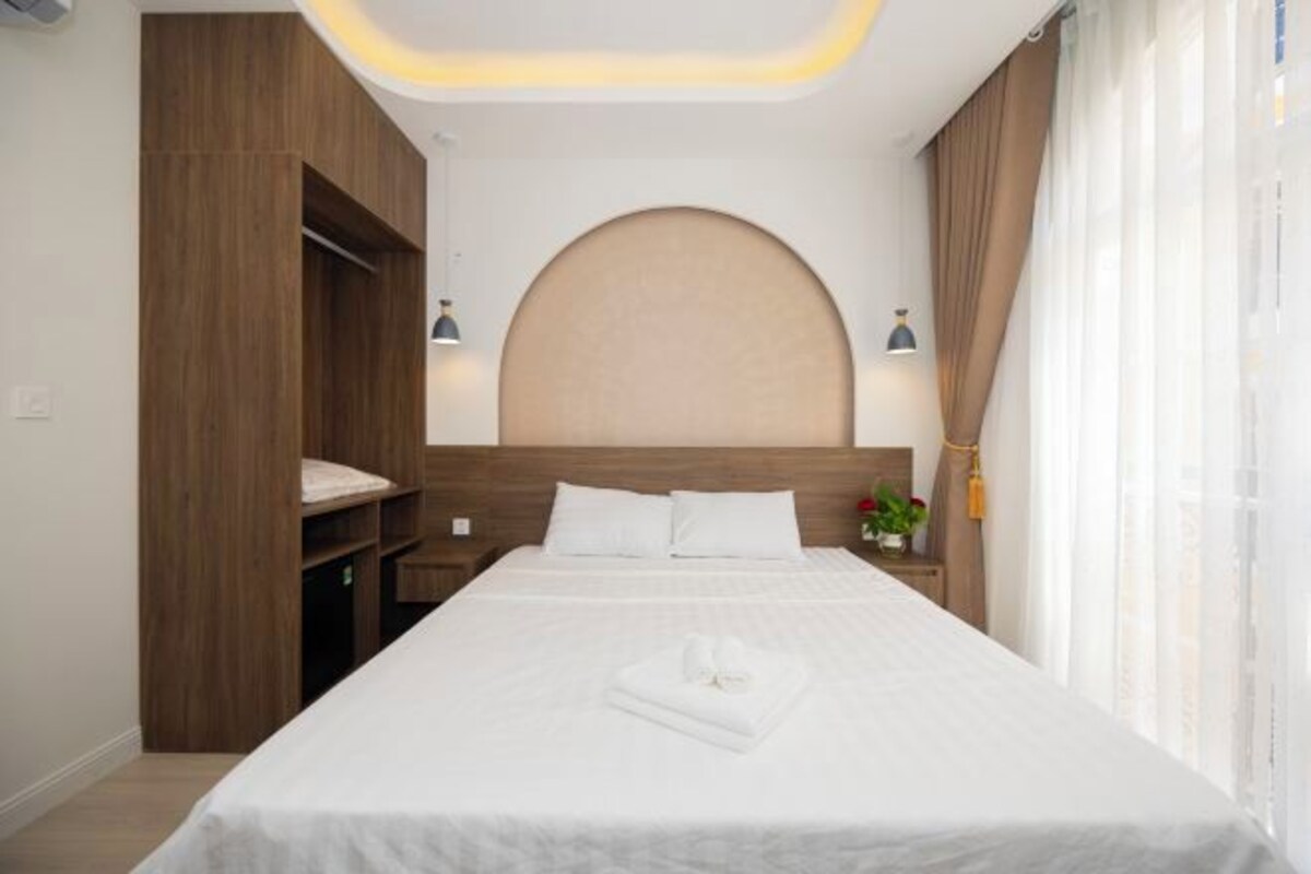 A hotel room with a large bed, a wooden wardrobe, a round headboard, and a nightstand with a lamp.