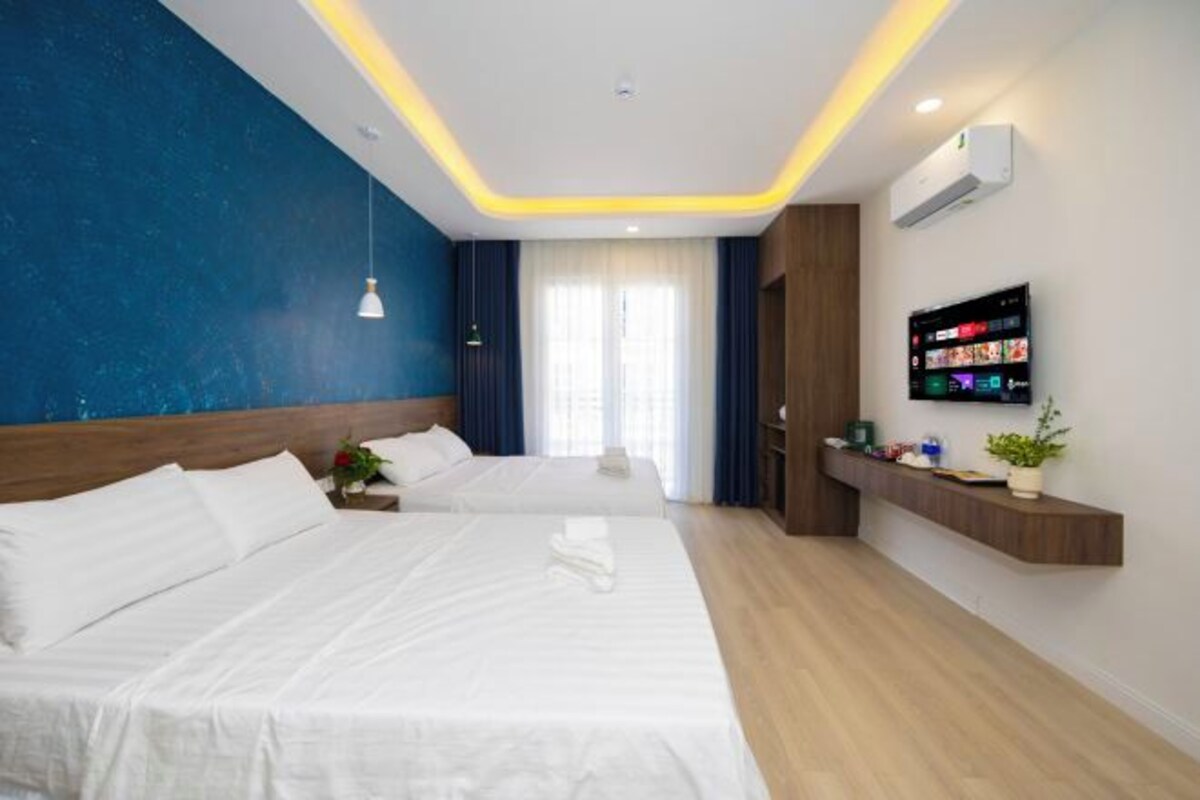A hotel room with two beds, a TV, a shelf with items, and a window with curtains.