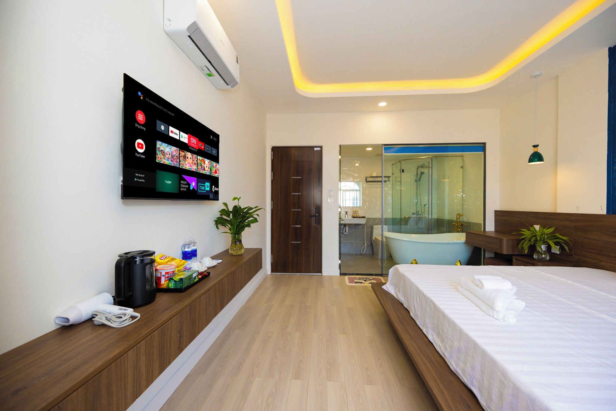 A modern hotel room with a flat-screen TV, a wooden bench with amenities, a large bed, and a bathroom visible through a glass door.