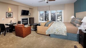 A hotel room with a bed, a desk, a television, a fireplace, and a large window with a view of buildings.