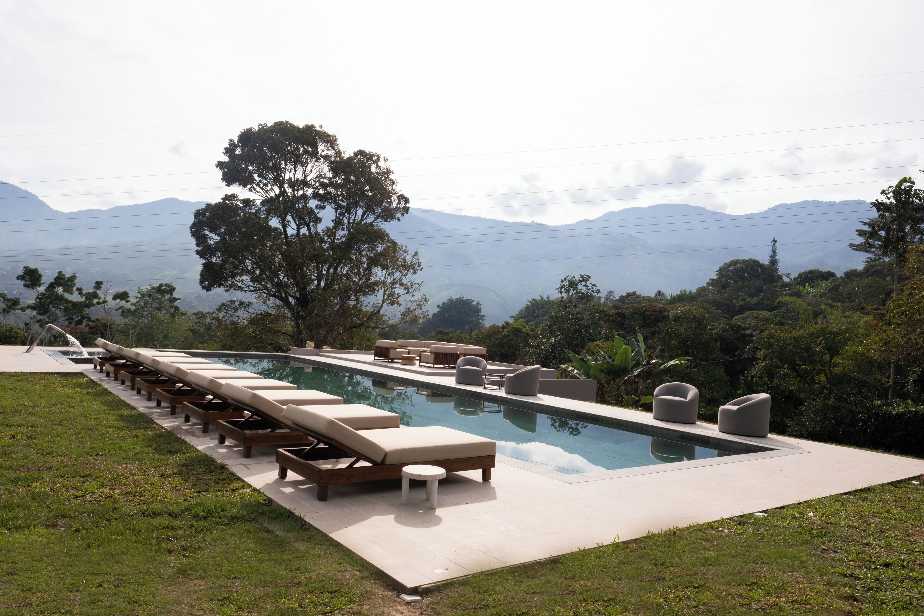 A swimming pool with lounge chairs and a table, surrounded by grass and trees, with mountains in the background.