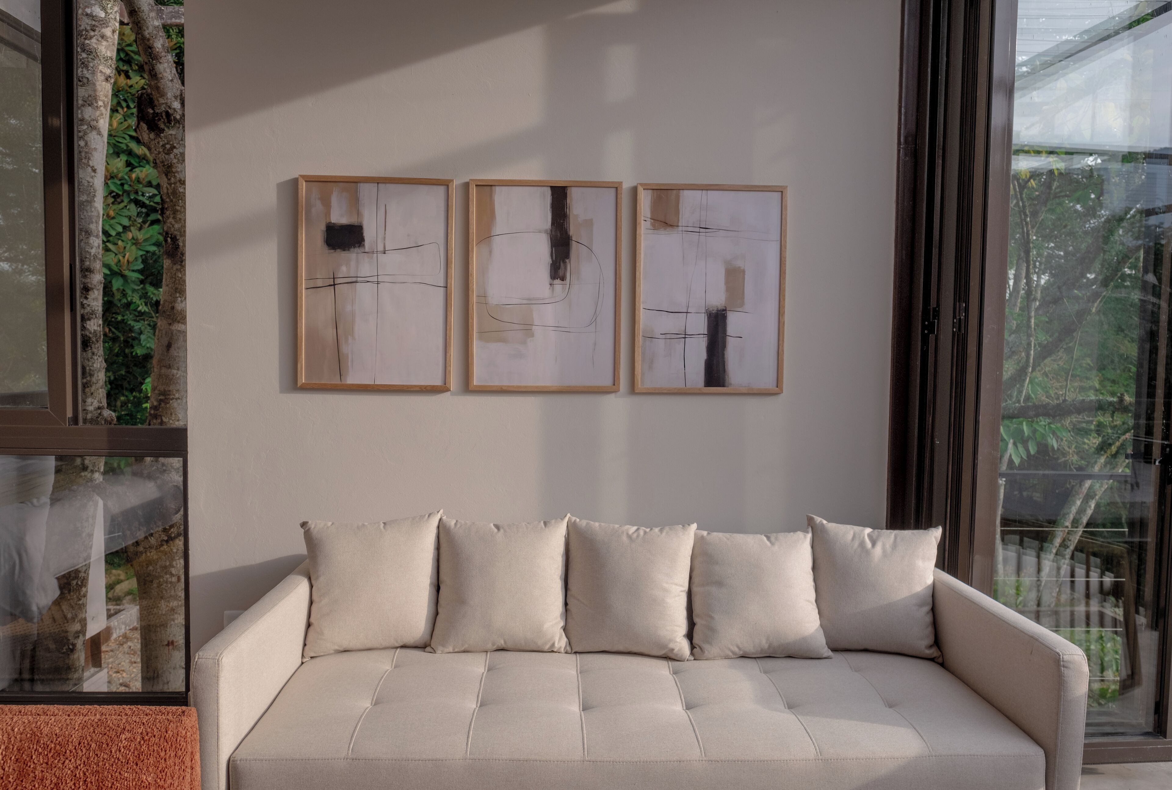 Uma sala de estar moderna com um sofá bege, três pinturas abstratas e uma grande janela com vista para o verde.