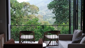A balcony with a view of lush greenery and mountains. There is a white sofa and two wooden chairs with cushions.