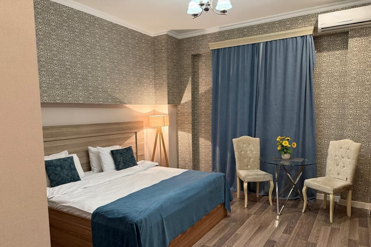 Une chambre d’hôtel avec un lit, deux chaises, une petite table ornée de fleurs, une lampe et un appareil de climatisation.
