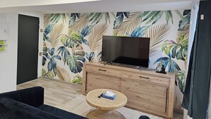 A living room with a flat-screen TV on a wooden cabinet, a dark sofa, a round wicker table, and a tropical leaf-patterned wall. - Cosy apartments Valenciennes center Train station and streetcar on foot (Valenciennes)