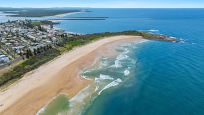 Vista aérea de un pueblo costero con playa de arena, aguas azules cristalinas y una zona residencial con casas y vegetación.