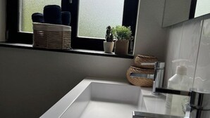 Une salle de bain avec un lavabo blanc, une fenêtre ornée de paniers remplis de serviettes et une petite plante en pot.