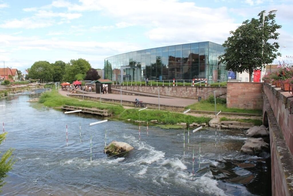 Un bâtiment moderne à la façade de verre, une rivière traversée par un pont en bois et un espace verdoyant où se trouvent quelques personnes.