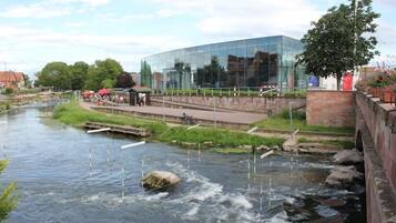 Un edificio moderno con facciata in vetro, un fiume con un ponte di legno e una zona erbosa con alcune persone.