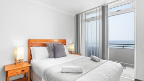 Habitación de hotel con una cama grande, dos mesitas de noche con lámparas, vista al mar desde un balcón y un cabecero de madera.