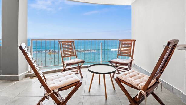 Un balcón con cuatro sillones de madera y una pequeña mesa redonda, con vista a una playa de aguas azules cristalinas y un litoral rocoso.