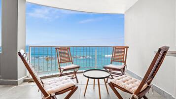 Un balcon doté de quatre fauteuils en bois et d’une petite table ronde, offrant une vue sur une plage aux eaux d’un bleu limpide et sur une côte rocheuse.