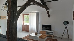En moderne stue med en stor træstamme, et fladskærms-tv på et hvidt stativ, en tripodlampe og et hyggeligt siddeområde med en sofa og sofabord.
