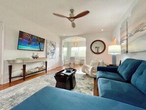 A living room with a blue sofa, a coffee table, and a ceiling fan. There is a painting on the wall and a lamp on a side table.