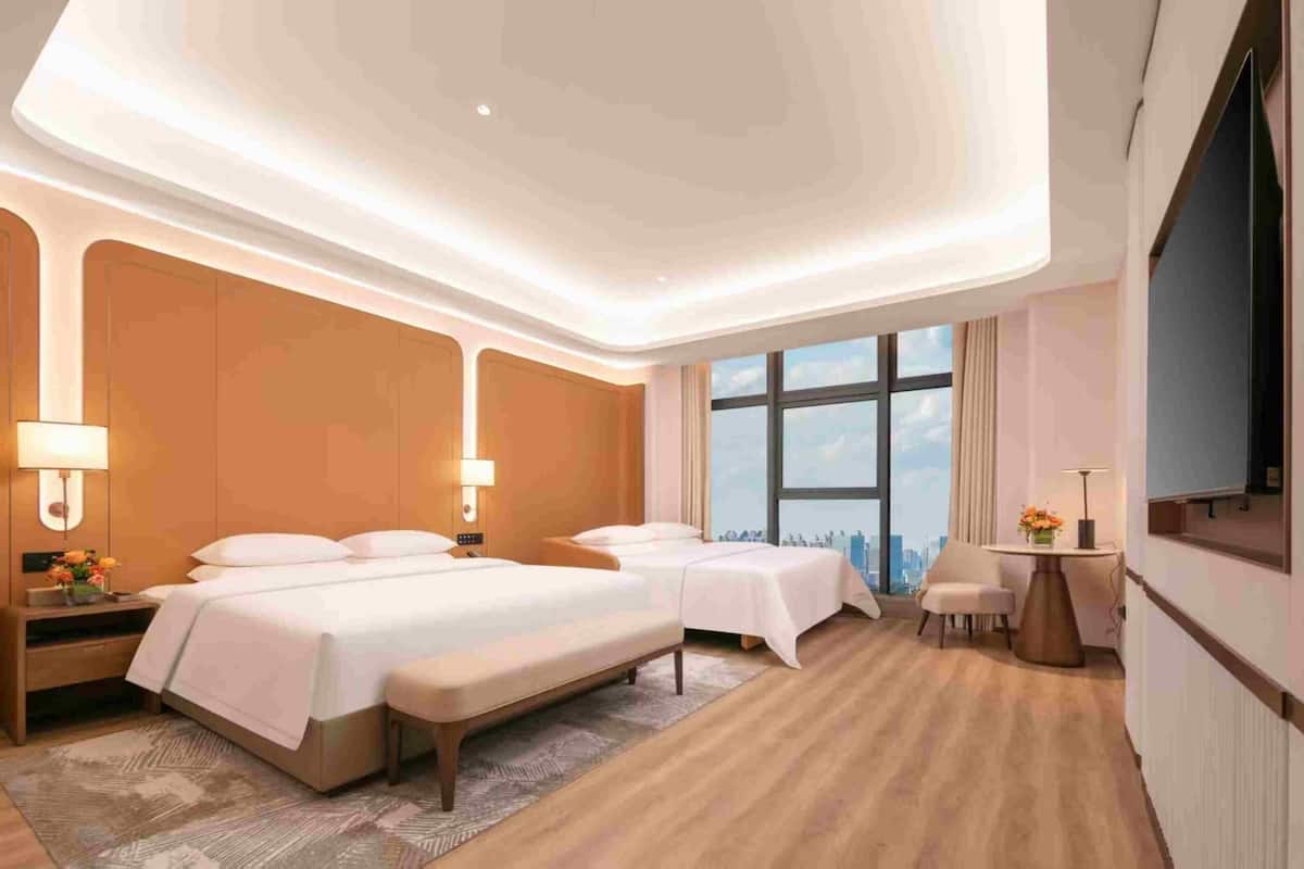 A hotel room with two beds, a desk, a chair, and a large window offering a city view.