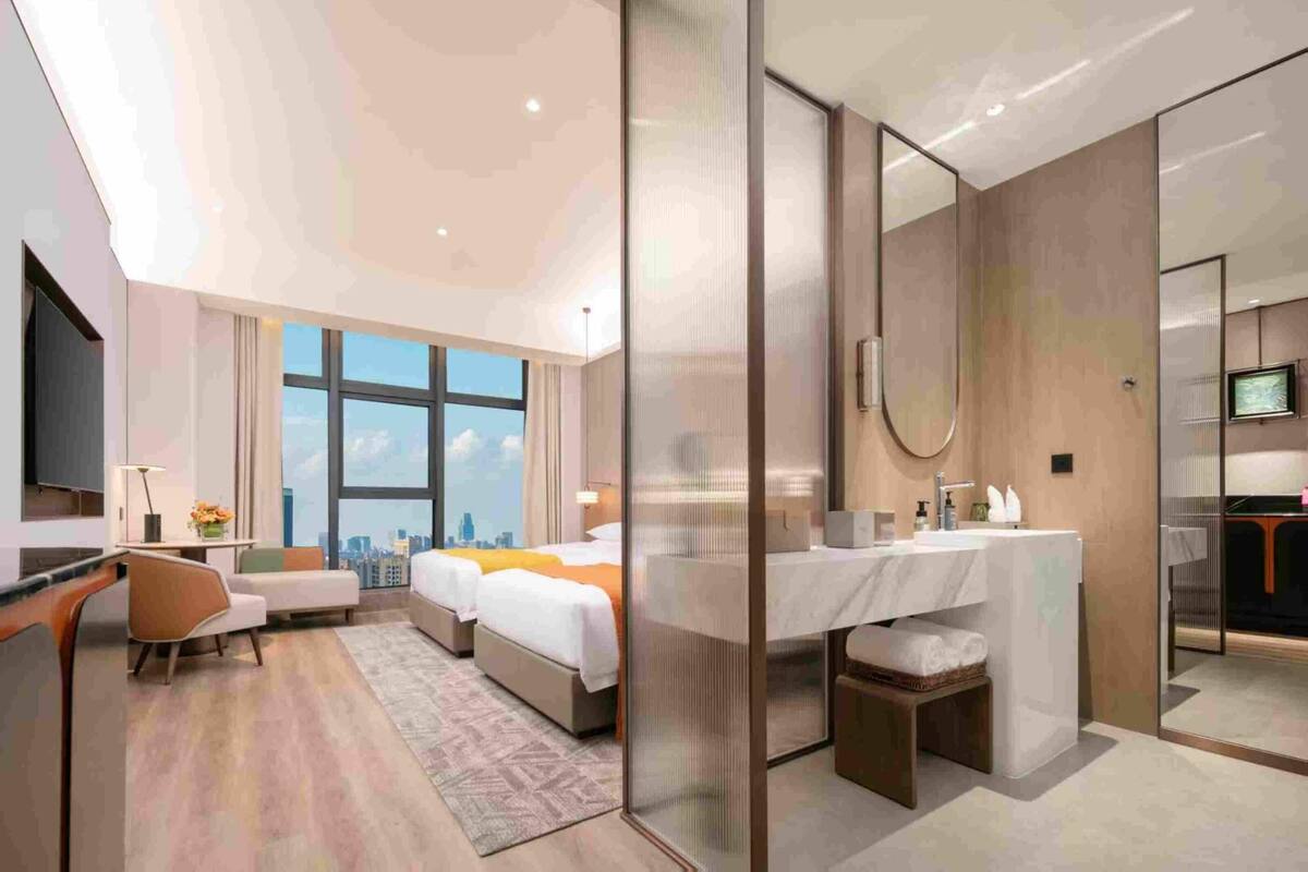 A modern hotel room with a large bed, a desk, a chair, a bathroom with a mirror, and a city view.