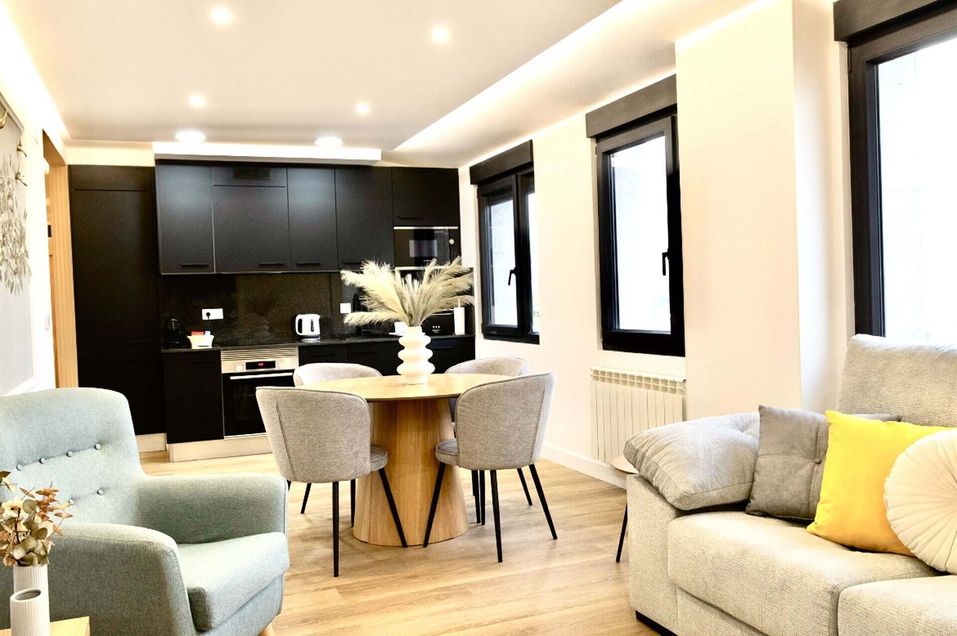 Un salon moderne avec un canapé gris, une table à manger en bois et quatre chaises, et une cuisine en arrière-plan, dotée de meubles sombres et d’une cuisinière.