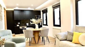 Una sala moderna con un sofá gris, una mesa de comedor de madera con cuatro sillas, y una cocina al fondo con armarios oscuros y una estufa.