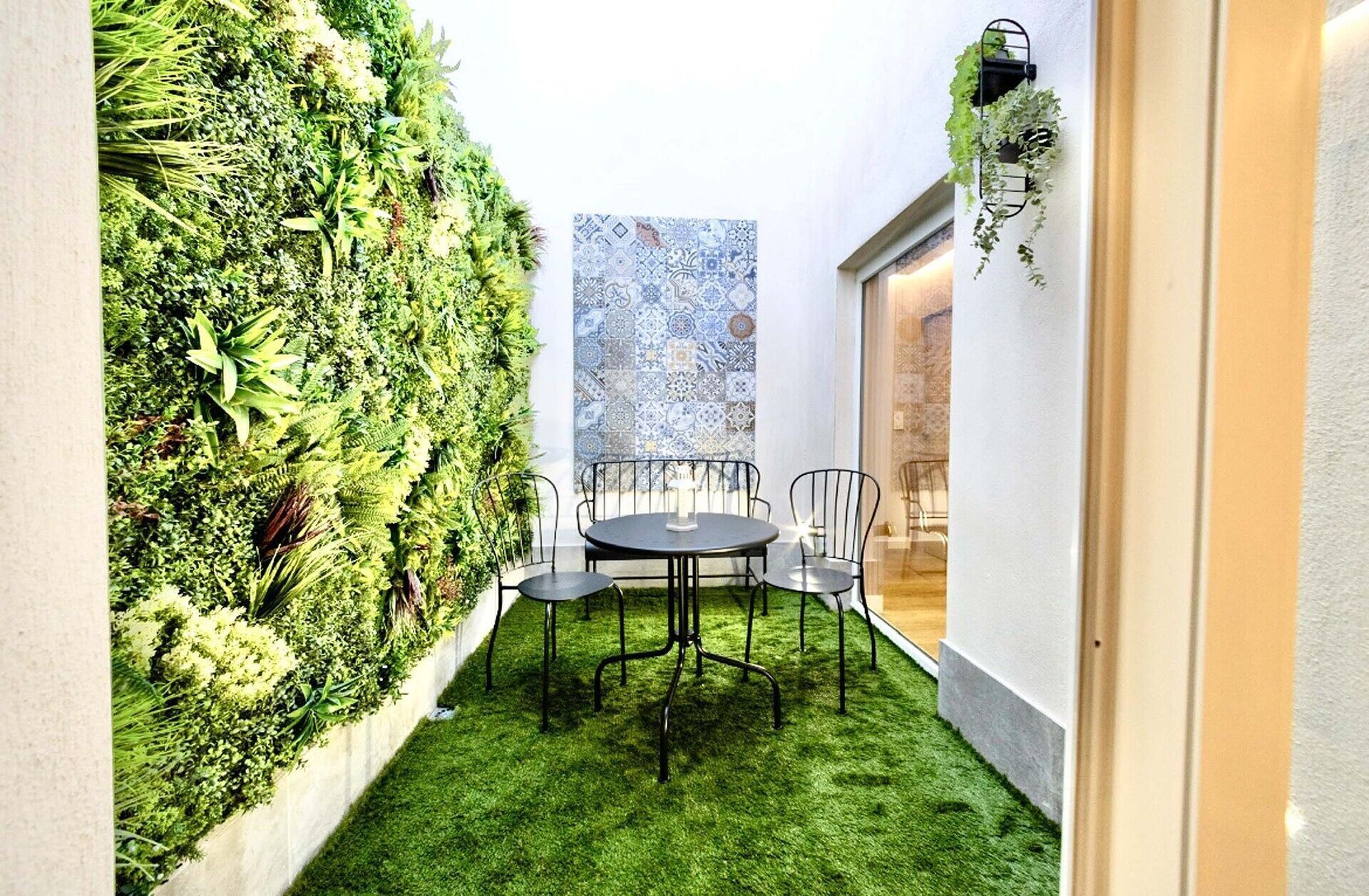 Un pequeño patio al aire libre con una mesa redonda y sillas, rodeado de abundante vegetación y un muro decorado al fondo.