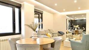 A modern living room with a round wooden table, grey upholstered chairs, a grey sofa with cushions, and a large window providing natural light.