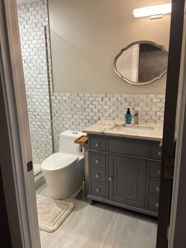 A bathroom with a white toilet, a grey vanity with drawers, a mirror, and a shower area with a tiled wall. - Charming 2-Bedroom Luxury Condo in the heart of Canton (Canton)