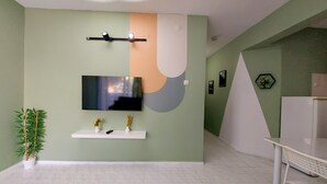 A modern living room with a flat-screen TV mounted on the wall, a white shelf with decorative items, and a potted plant. - Mar Soleil Apartments (Marmaris)