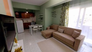 A living room with a beige sofa, a dining table with chairs, a kitchen area with cabinets and a refrigerator, and a large window with curtains. - Mar Soleil Apartments (Marmaris)