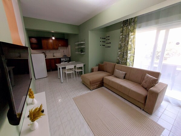 A living room with a beige sofa, a dining table with chairs, a kitchen area with cabinets and a refrigerator, and a large window with curtains.