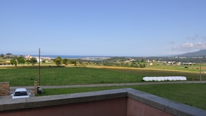 A view from a balcony overlooking a green field with hay bales, a road, and distant buildings.