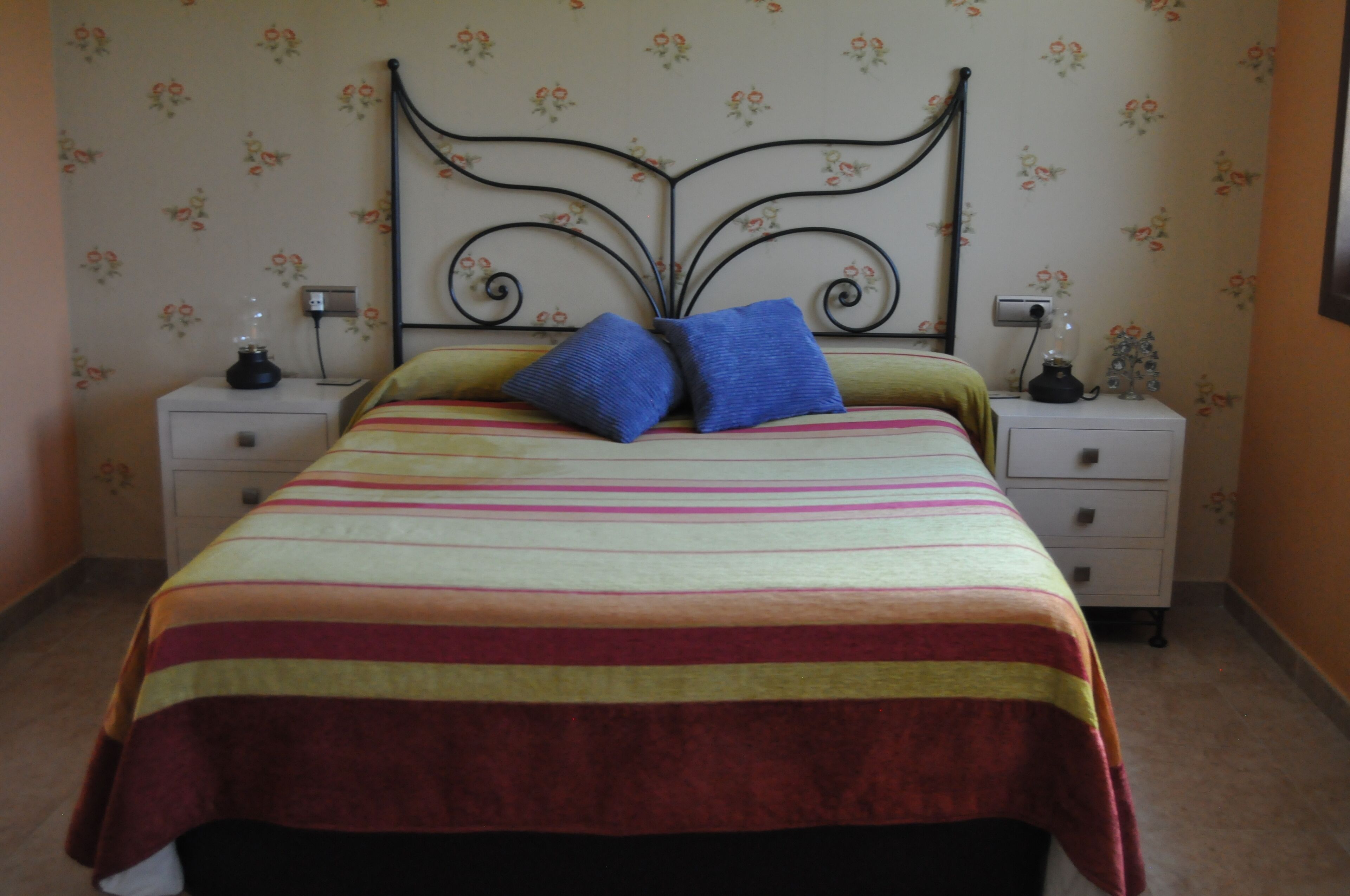 A bed with a striped bedspread, two pillows, and a metal headboard. There are two bedside tables with lamps and a wall with floral wallpaper.