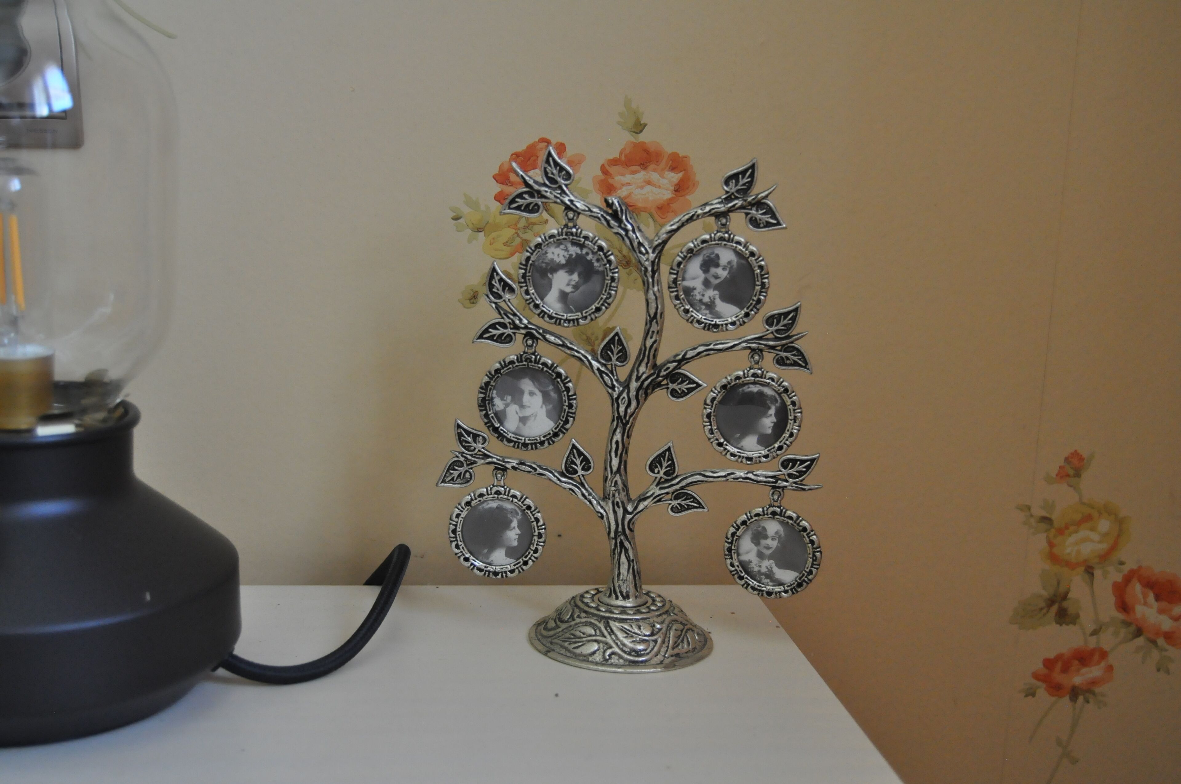 Un arbre décoratif en métal, orné de cadres photo circulaires sur ses branches, est placé sur une table devant un mur recouvert d’un papier peint à motifs floraux.